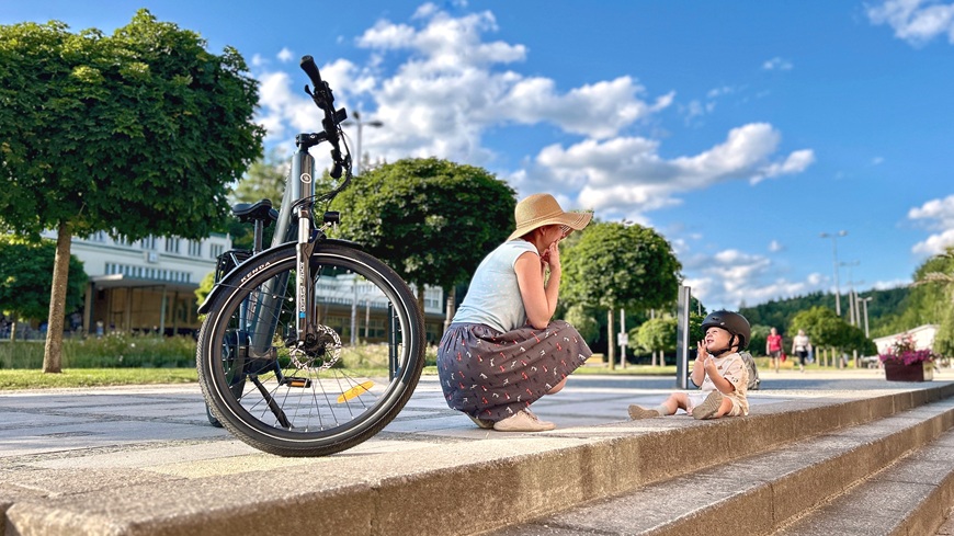 Recenzia: Elektrobicykel GoGEN Aspen MX 720 sa postará o to, aby vaše cyklistické túžby nič nebrzdilo Elektrobike GoGEN Aspen MX 720 v šedo-čiernom prevedení, opretý o stojan, na pozadí žena s batoľaťom