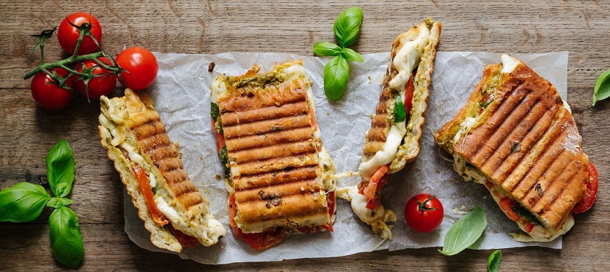 ETA Sorento: 18 tipov na školské desiaty, ktoré si deti zamilujú Grilované panini s mriežkou plnené mozzarellou, paradajkami a bazalkovým pestom, nakrájané na papieri na pečenie na drevenom stole. Príprava v sendvičovači ETA Sorento.