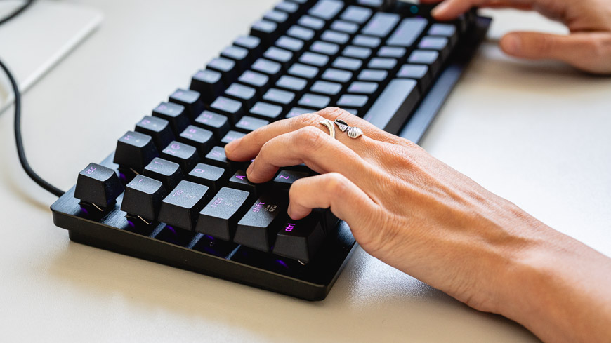 Herná klávesnica Razer Huntsman Tenkeyless V3 8KHz s fialovým podsvietením leží na stole, na klávesnici sú položené ruky, pohľad zboku