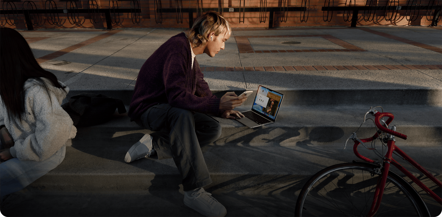 Osoba sedí vonku na betónových schodoch, má nasadený jeden AirPod, drží iPhone a pomocou trackpadu ovláda Mac položený na zemi pred sebou, neďaleko leží červený bicykel a sedí druhá osoba